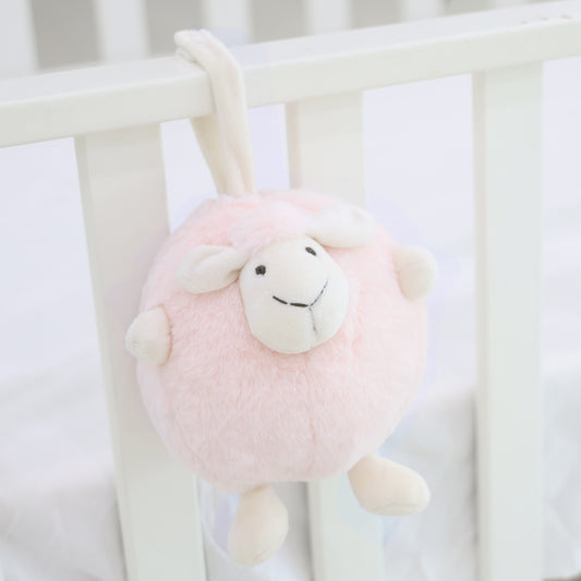 A round and plush sheep toy in soft pink by Le Petit Society hangs off the frame of a baby cot.