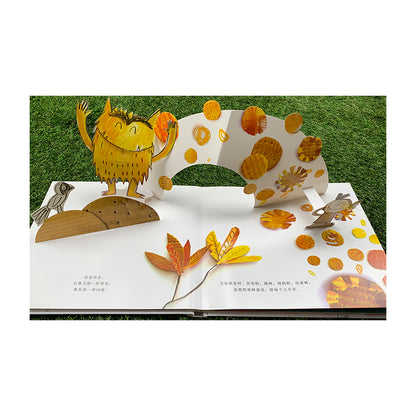 Colorful pop-up book on grass. Left page shows a smiling yellow monster and a bird on a hill. Center has a white arch with orange-yellow circles. Right page shows a girl and orange leaves, with scattered circular patterns.