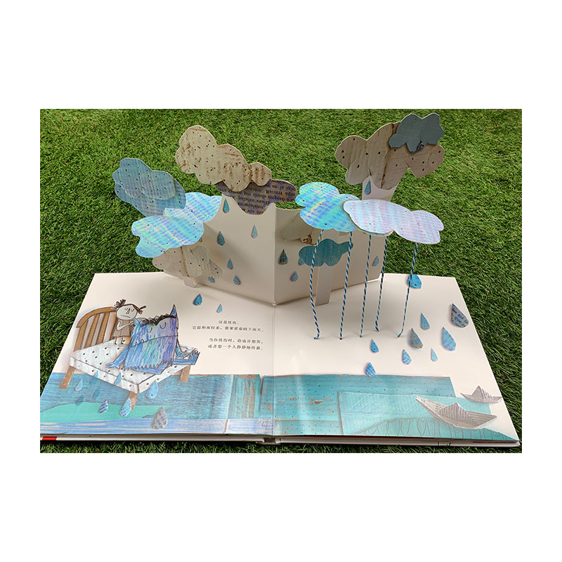 Pop-up book on grass showing a rainy scene with blue water and paper boats. A small girl and a colorful monster sit on a bed. Multiple clouds hang above with strings of raindrops falling down onto the scene. Text is visible beneath the pop-up.