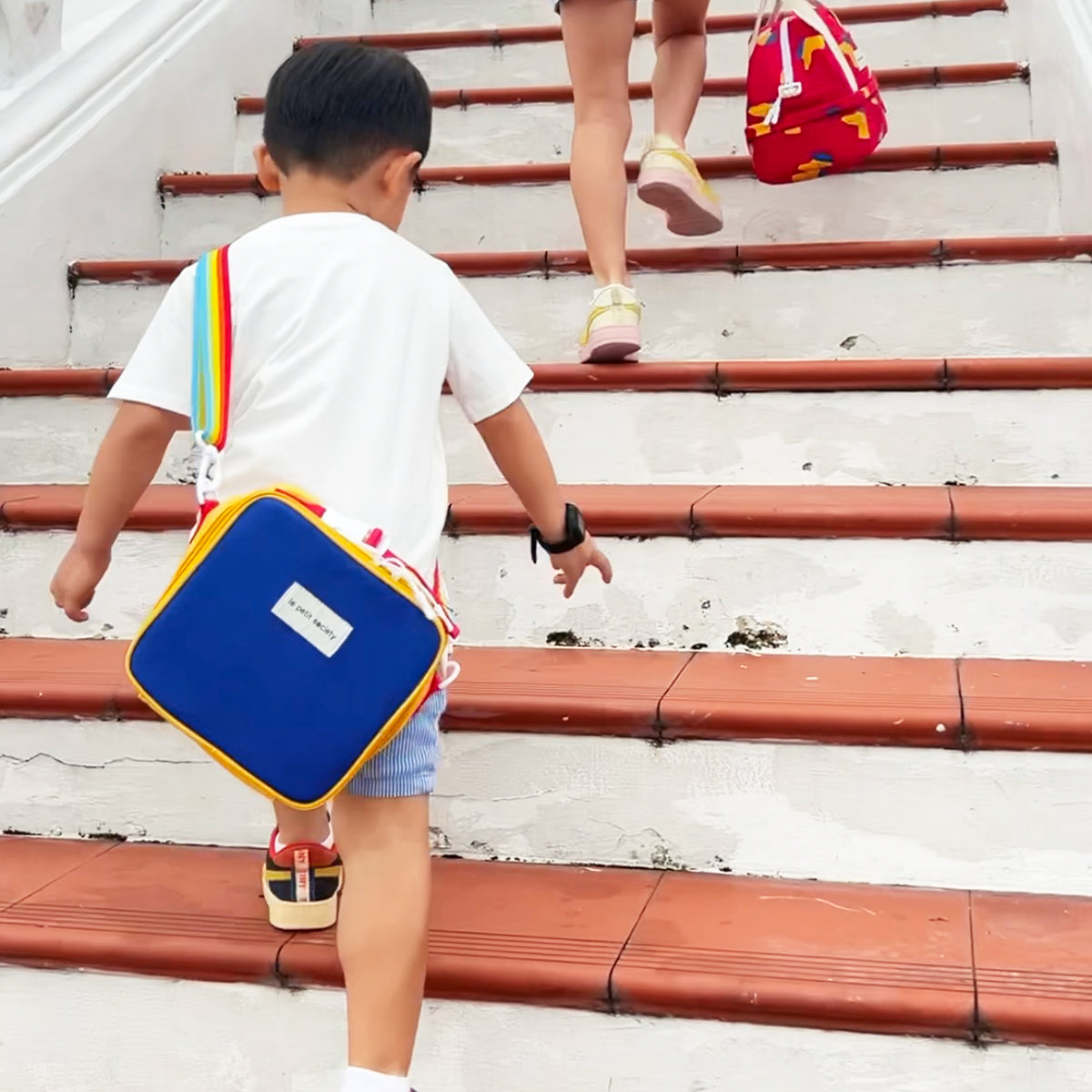 A preschool kid carrying Le Petit Society's thermal lunch bag with a colour block design and a rainbow strap.
