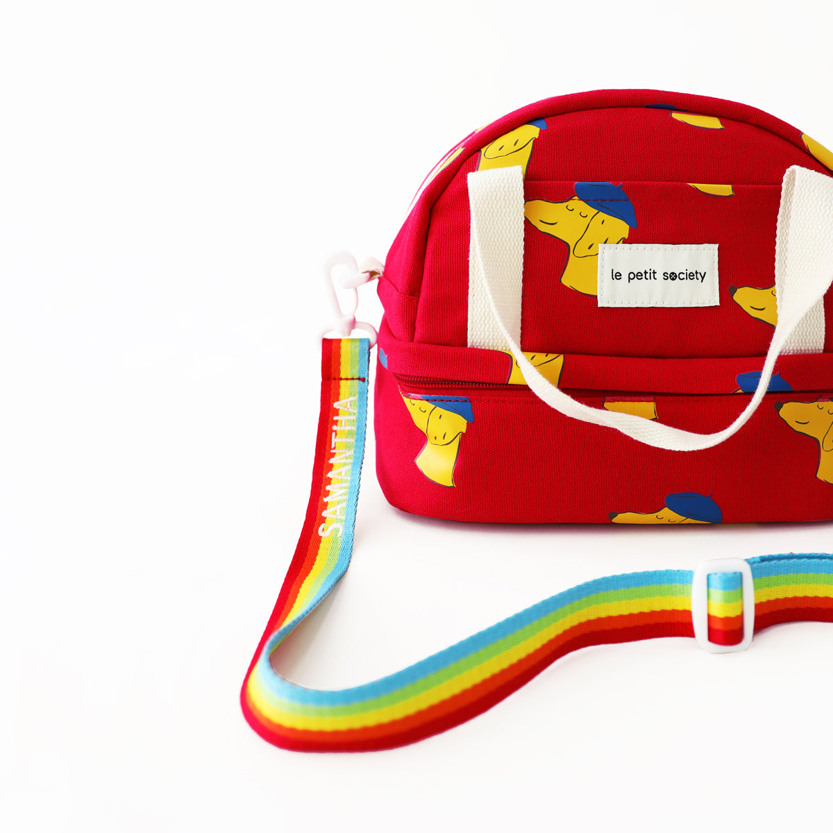 A red kid's thermal lunch bag with a dog print and a rainbow strap from Le Petit Society.