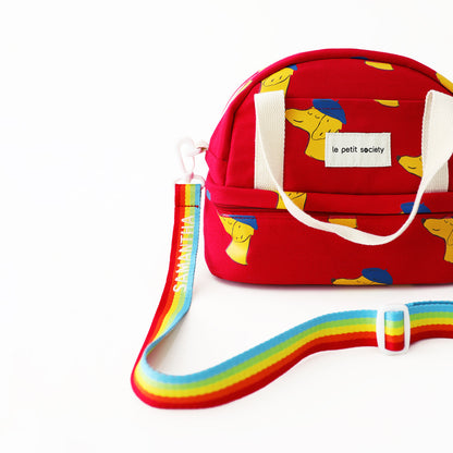 A red kid's thermal lunch bag with a dog print and a rainbow strap from Le Petit Society.