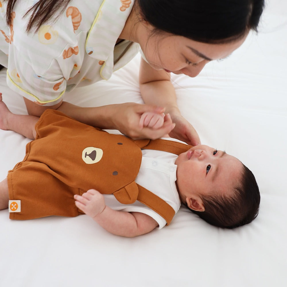 An animal-themed baby romper by Le Petit Society, featuring a brown bear's face at the front, complete with brown bottoms and brown cross back straps over a white base.