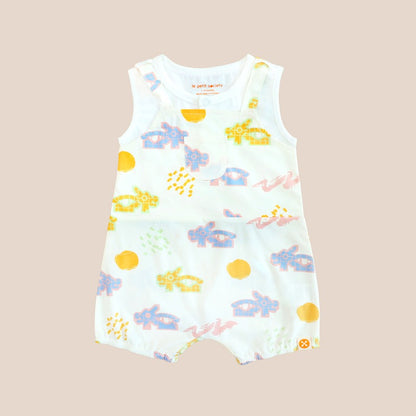 White baby overall from Le Petit Society with colorful dragon playground and sun print in pastel blue, yellow, green, and pink, featuring sleeveless straps and a front pocket.