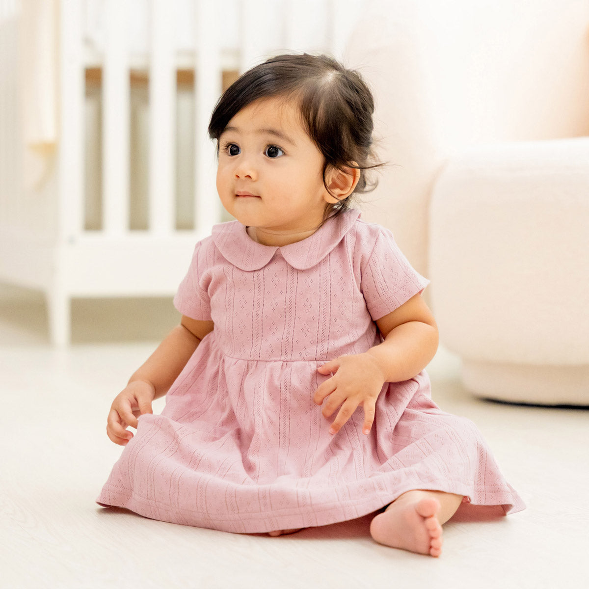 A baby girl wearing a plum-coloured, pointelle cotton dress with a peter pan collar from Le Petit Society.