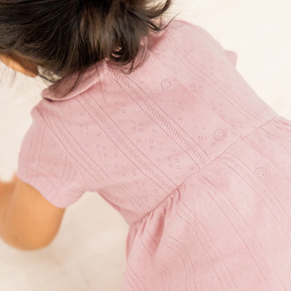 A baby girl wearing a plum-coloured, pointelle cotton dress with a peter pan collar from Le Petit Society. It also comes with snap buttons at the back for easy changing.