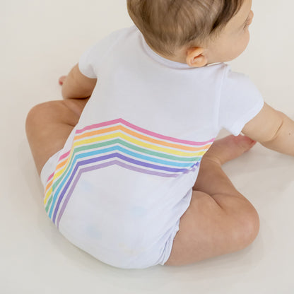 A baby in a white romper with a pastel print of the right arc of a rainbow by Le Petit Society.