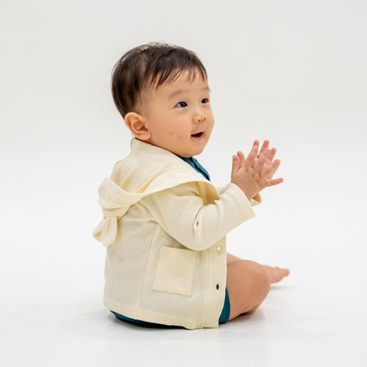 A baby boy wearing a bear cardigan in cream waffle jersey, which comes with buttons and pockets at the front, as well as a hood with little bear ears.