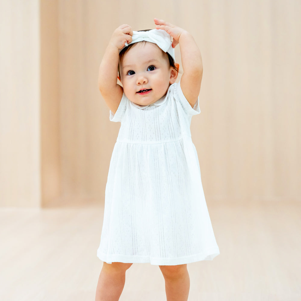 A baby girl wearing a peter pan collar dress in white pointelle cotton with a matching bow headband.