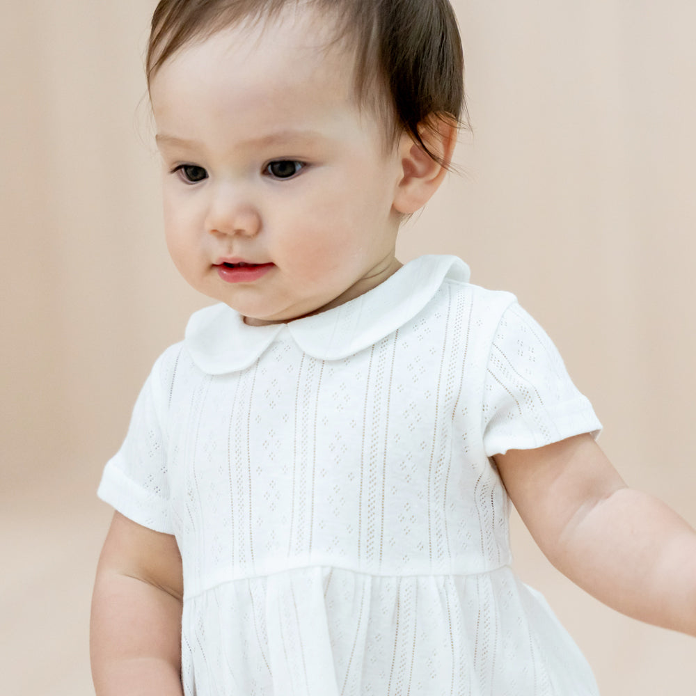 A baby girl wearing a peter pan collar dress in white pointelle cotton.