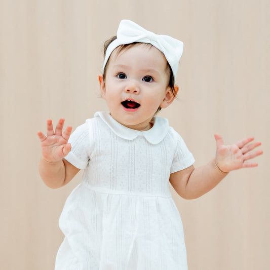 A baby girl wearing a matching outfit from Le Petit Society, consisting of a peter pan collar dress and bow headband in white pointelle cotton.