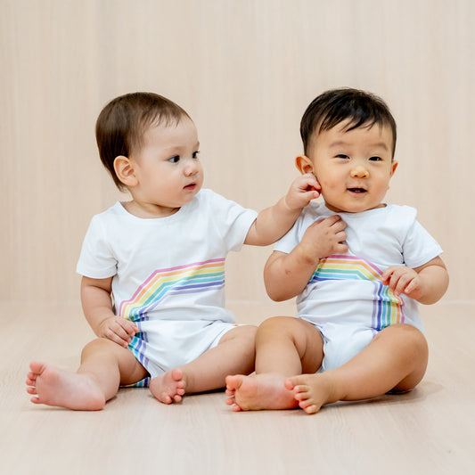 Two babies wearing white rompers with a print of the left and right arc of a rainbow respectively, a matching outfit that creates a complete rainbow when placed side-by-side.