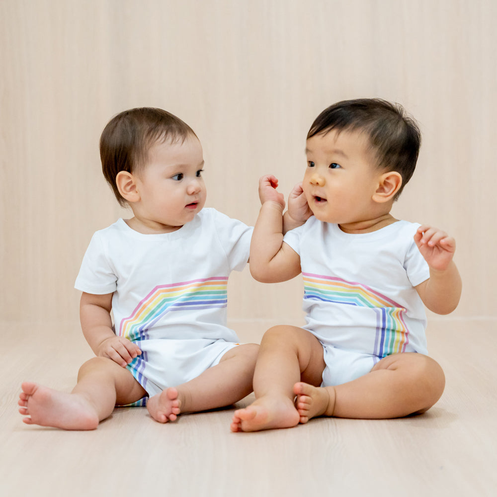 Two babies wearing white rompers with a print of the left and right arc of a rainbow respectively, a matching outfit that creates a complete rainbow when placed side-by-side.