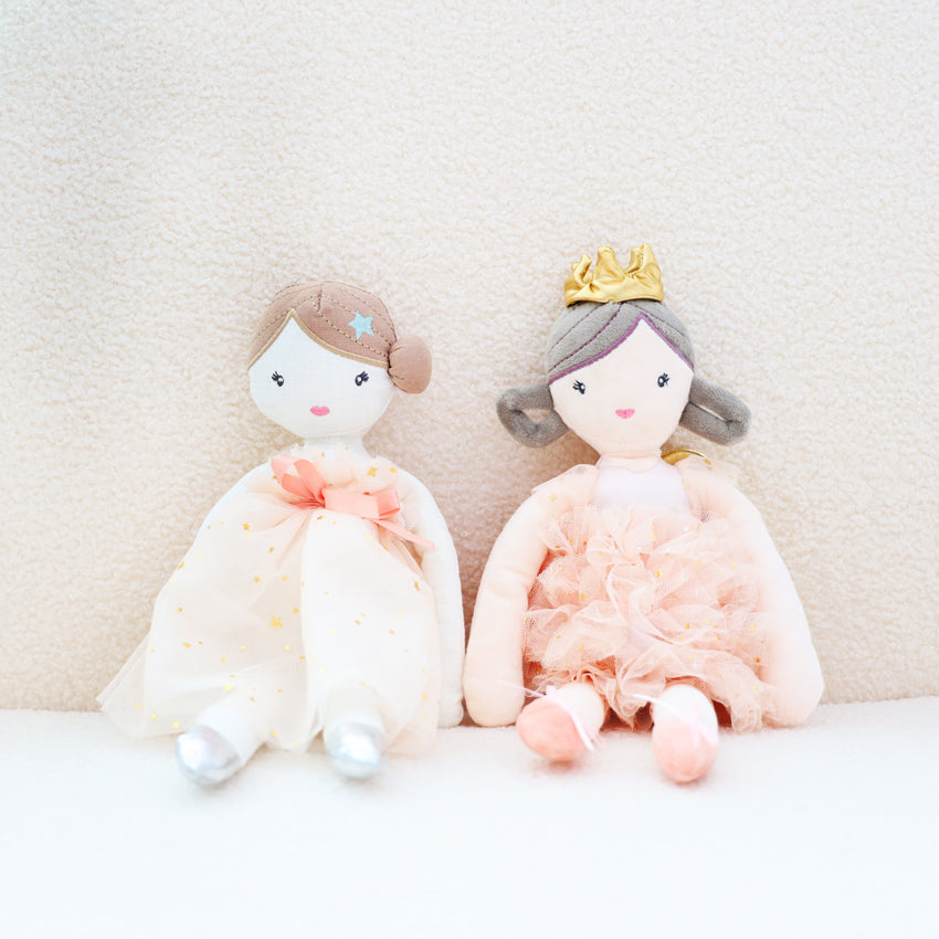 A pair of plush dolls by Le Petit Society are sitting together against a sofa. On the left is a ballerina doll in a pink dress and silver ballet shoes, and on the right, is a princess doll in a pink dress and golden crown.