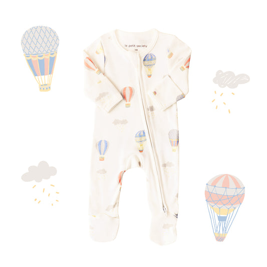 An organic baby zip sleepsuit from Le Petit Society. It is beige in colour with a print of hot air balloons and foldable mittens and footies.