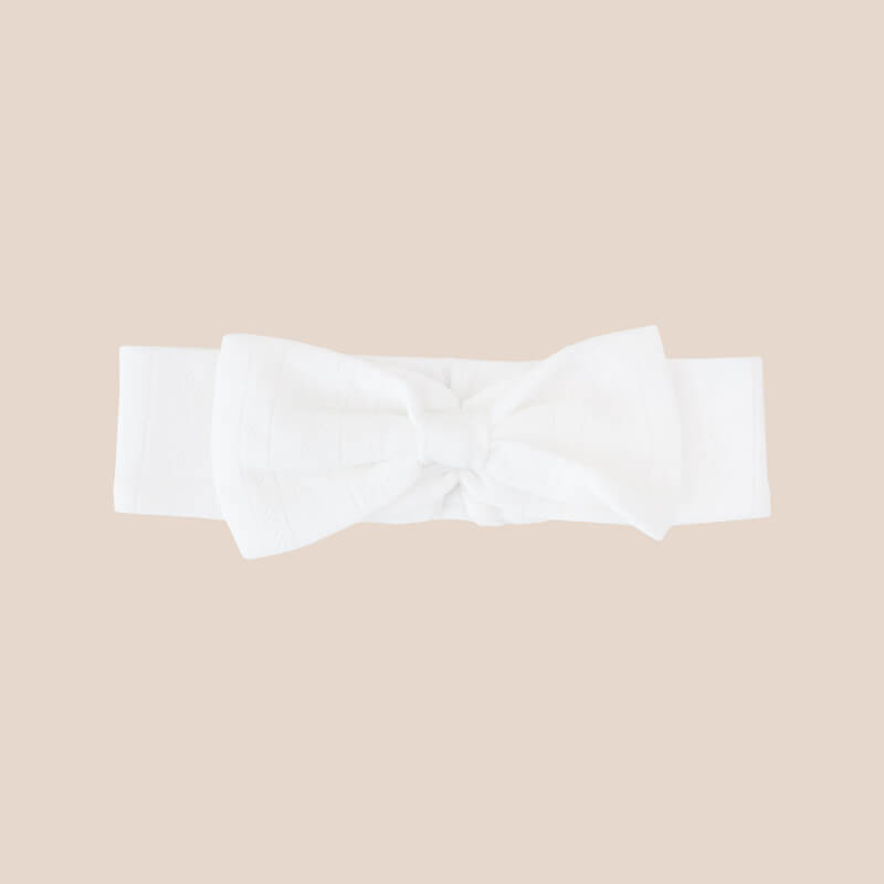A baby headband from Le Petit Society. It is made from a white pointelle cotton fabric and comes with a large bow in the middle.