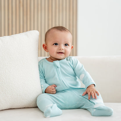 A smiling baby wearing an organic zip sleepsuit with foldable mittens and footies by Le Petit Society, featuring a print of clouds over a teal base. 