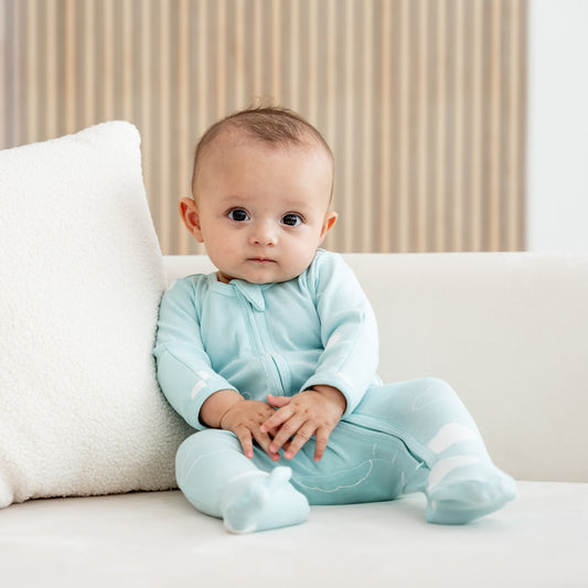 A baby wearing an organic zip sleepsuit with foldable mittens and footies by Le Petit Society, featuring a print of clouds over a teal base. 