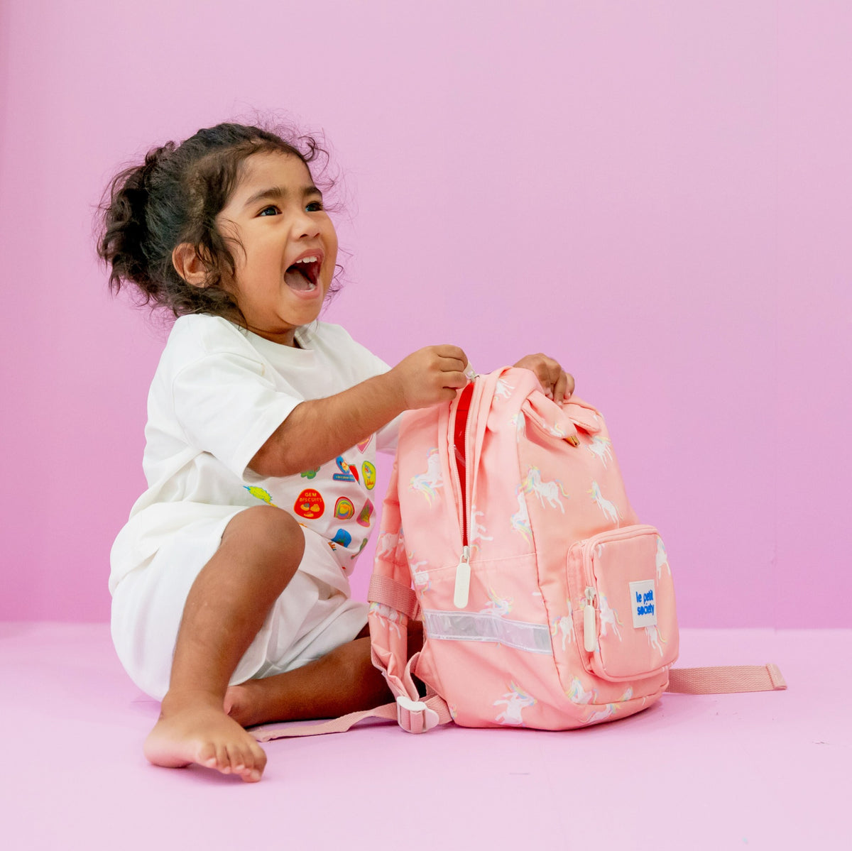 A toddler girl smiling with a pink unicorn print preschool bag.