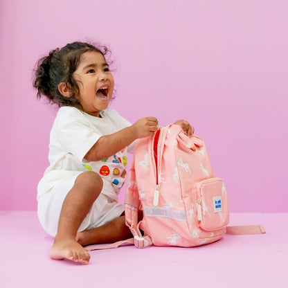 A toddler girl smiling with a pink unicorn print preschool bag.