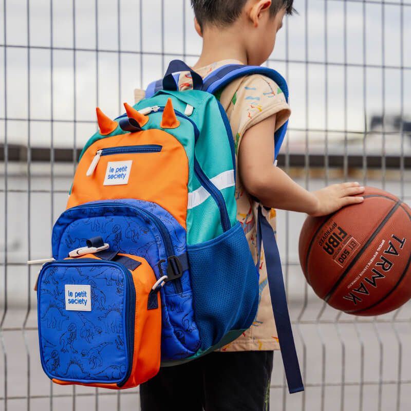A primary school boy playing basketball while carrying the single pocket school bag and lunch bag bundle.