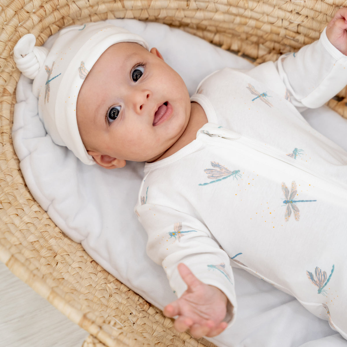 A baby wearing a matching hat and zip sleepsuit in dragonfly print by Le Petit Society.