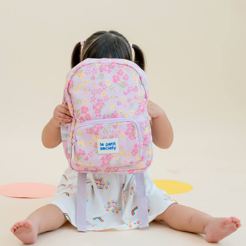 A toddler girl hiding behind a purple, floral-print preschool bag.