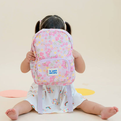 A toddler girl hiding behind a purple, floral-print preschool bag.