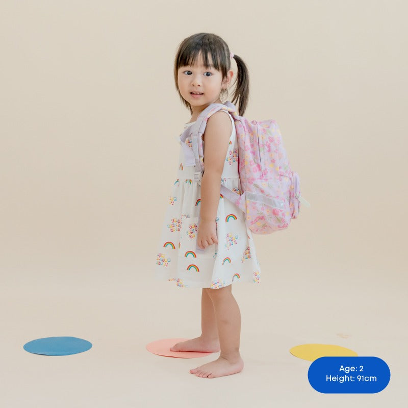A toddler carrying a purple, floral-print preschool bag.