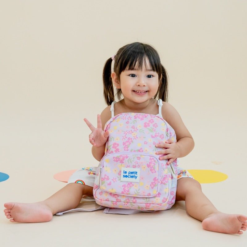 A toddler smiling with a purple, floral-print preschool bag.