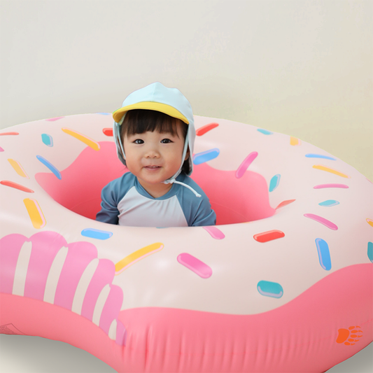 Baby wearing Le Petit Society Baby Penguin Sunhat in light blue with yellow visor, sitting inside a pink donut inflatable pool float in matching penguin rash guard swimsuit