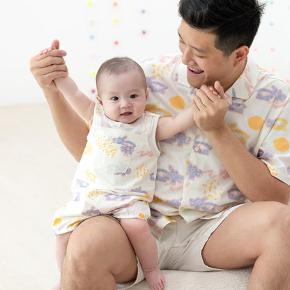 White baby overall from Le Petit Society with colorful dragon playground and sun print in pastel blue, yellow, green, and pink, featuring sleeveless straps and a front pocket.
