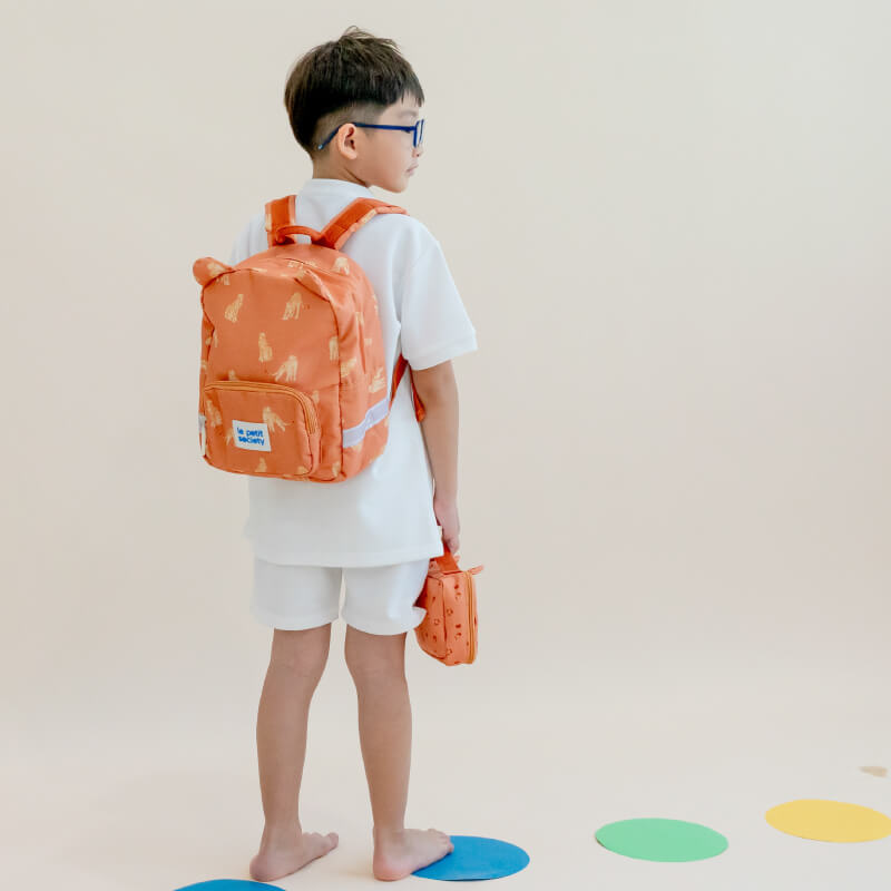 A primary school boy carrying an orange school bag and pencil case in tiger design. 

