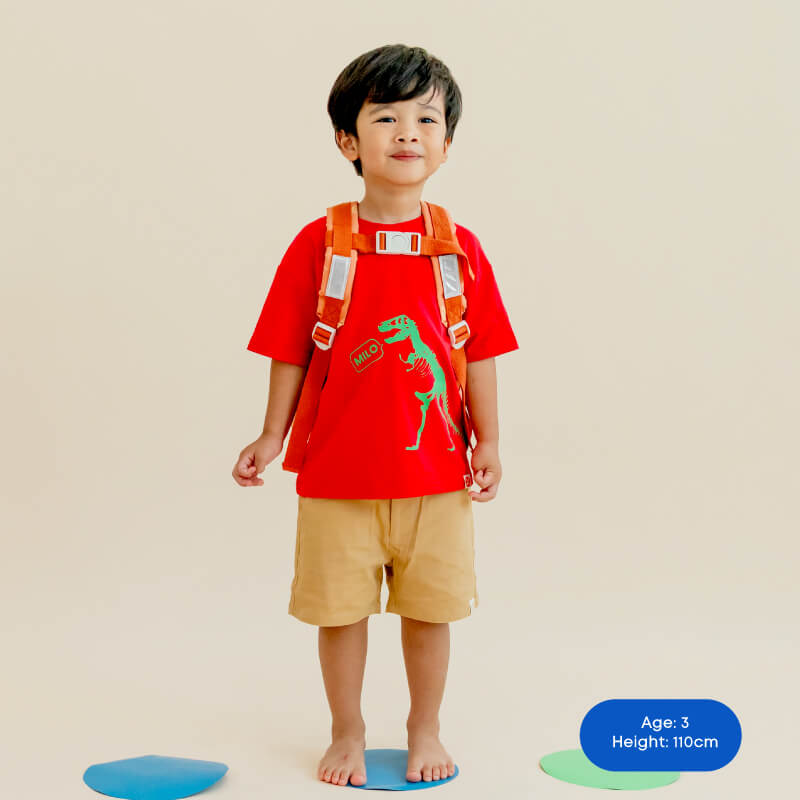A preschool boy in Le Petit Society's red shirt with dinosaur print and an orange school bag.