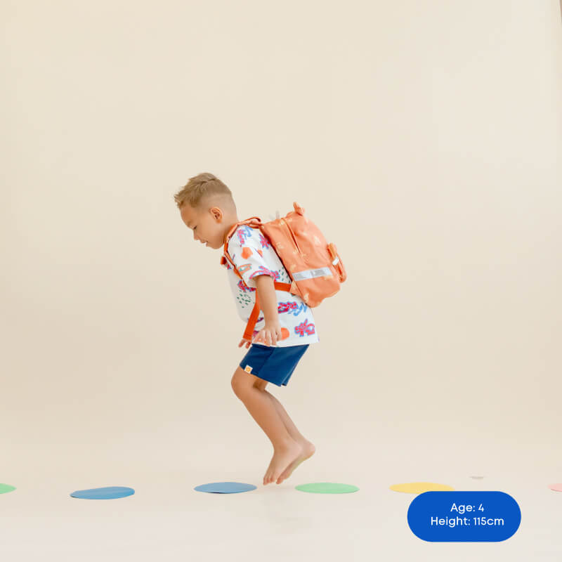 A little boy light on his feet and jumping while carrying an orange preschool bag.