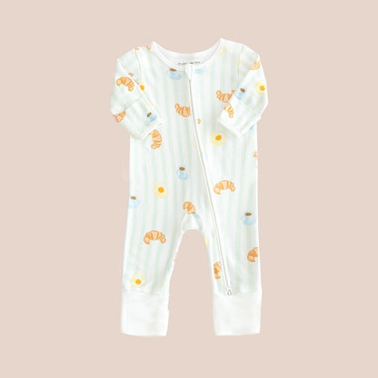 A baby bamboo sleepsuit with green stripes and a print of breakfast food: croissants, coffee and sunny side-up eggs.