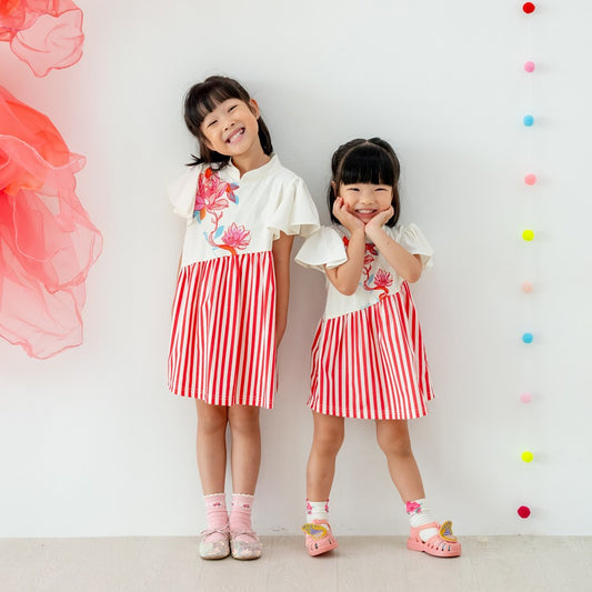 Two young girls wearing matching cream lotus-print tops paired with red-and-white striped skirt dresses from the Le Petit Society x Sheila Sim CNY 2026 collection stand smiling against a bright backdrop decorated with coral organza fabric and colourful po