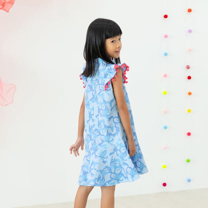 A young girl wearing a light blue ruffle cheongsam dress with pink pom-pom trim from the Le Petit Society x Sheila Sim CNY 2026 collection turns slightly to look back over her shoulder. She stands against a bright backdrop decorated with colourful hanging