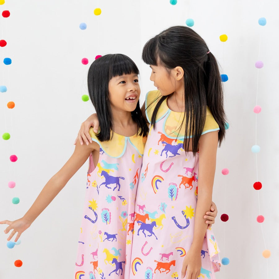 Two girls stand close together in matching Rainbow Horse dresses, smiling and chatting as they hold each other in a warm side hug. The bright, multicoloured horse motifs and the yellow scalloped collars give the outfits a fun, festive charm, while the rai
