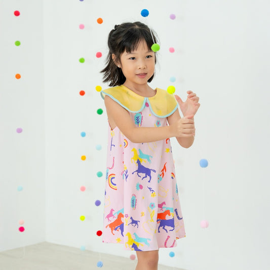 A young girl wears the Rainbow Horse dress from the Le Petit Society x Sheila Sim CNY 2026 Collection. The pastel pink dress features colourful horses, rainbows and lucky icons, paired with a soft yellow scalloped collar. She reaches for the hanging pom-p