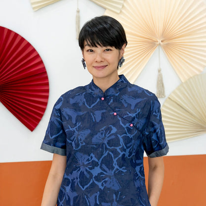 A female model wearing a navy blue chambray floral cheongsam-inspired dress from the Le Petit Society x Sheila Sim CNY 2026 collection stands in front of a two-tone orange and white wall decorated with red and cream paper fans. She smiles softly toward th