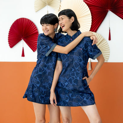 Two female models wearing matching navy blue chambray floral cheongsam-inspired dresses from the Le Petit Society x Sheila Sim CNY 2026 collection stand together, smiling with their arms around each other. They are posed in front of a two-tone orange and 