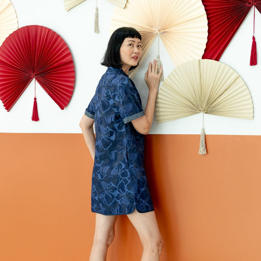 A female model wearing a navy blue chambray floral cheongsam-inspired dress from the Le Petit Society x Sheila Sim CNY 2026 collection stands sideways in front of a two-tone orange and white wall decorated with red and cream paper fans. She looks back tow