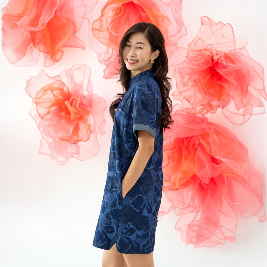 A female model wearing a navy blue chambray floral dress from the Le Petit Society x Sheila Sim CNY 2026 collection stands sideways with her hands in her pockets, smiling. She is posed in front of large, soft coral-pink organza flower decorations on a whi