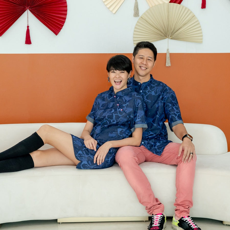 A female model wearing a navy blue chambray floral cheongsam-inspired dress sits leaning comfortably against a male model dressed in a matching chambray shirt from the Le Petit Society x Sheila Sim CNY 2026 collection. They sit on a white sofa in front of