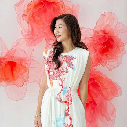 A female model wearing a cream dress with vibrant pink and blue lotus floral prints from the Le Petit Society x Sheila Sim CNY 2026 collection looks to the side while standing against a backdrop of large coral organza flowers.