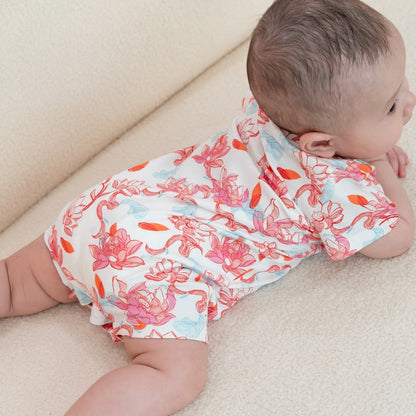 A baby lies comfortably on a soft cream surface, wearing the lotus-print romper from the Le Petit Society x Sheila Sim CNY 2026 collection. The back view shows off the full lotus artwork in flowing corals, pinks, and touches of blue — a gentle, dreamy pri
