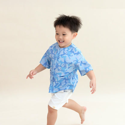 A young boy wearing a light blue printed short-sleeve shirt and white shorts from the Le Petit Society x Sheila Sim CNY 2026 collection runs joyfully across a soft cream background, smiling with his arms outstretched.