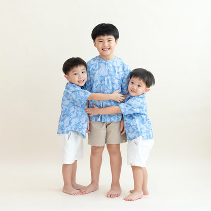 Three young boys wearing matching light blue printed shirts from the Le Petit Society x Sheila Sim CNY 2026 collection stand together in a group hug against a soft cream background. The older boy is in the center wearing beige shorts, while the two younge