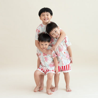 Three boys in the matching Lotus Collection outfits stand close together, hugging and smiling. The coordinated shirts and bold striped shorts create a bright, cheerful palette, while their warm interaction captures the sweetness of family bonding during C